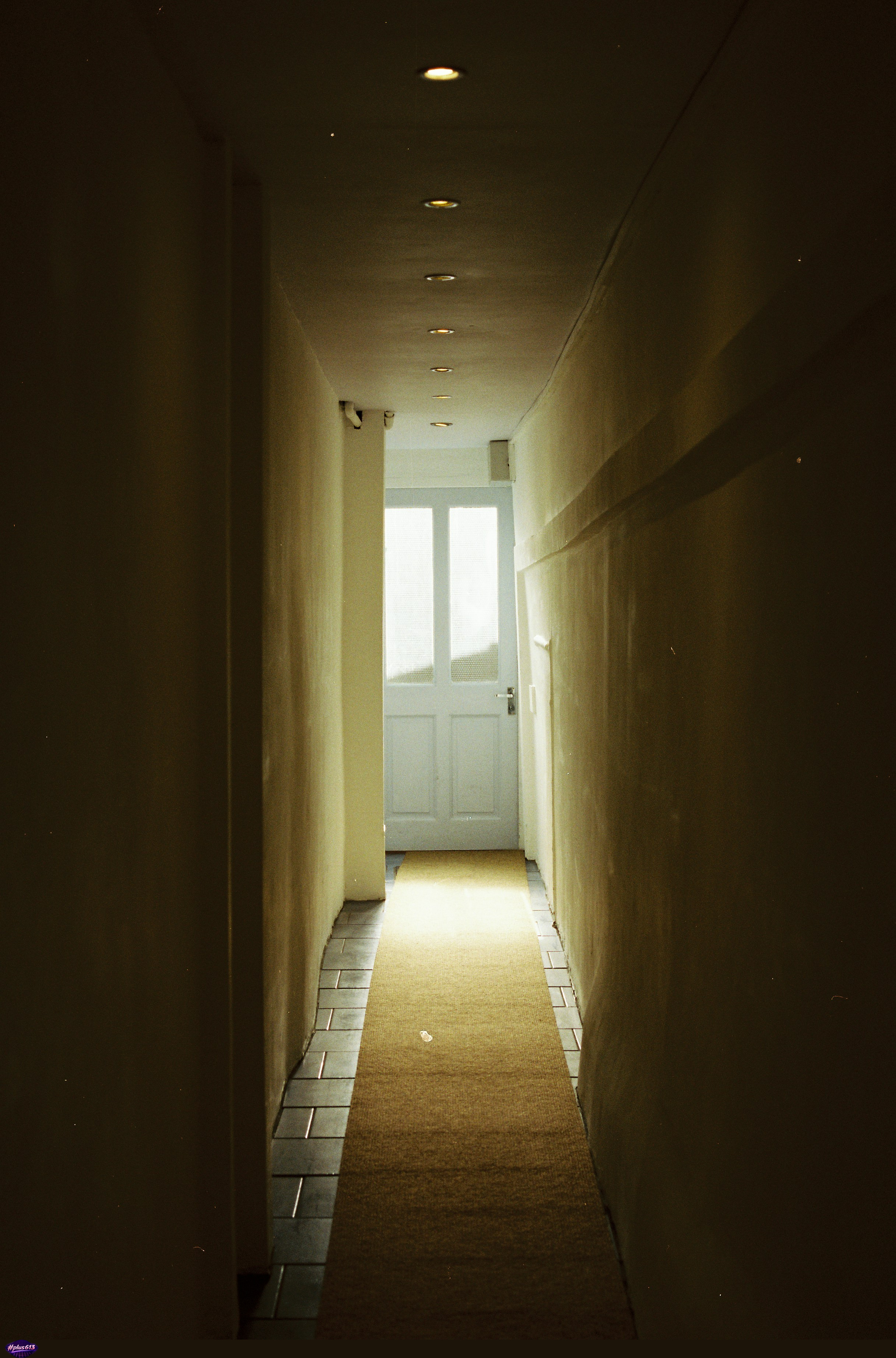 Long, dark hallway leading to a bright white door by Barney Goodman