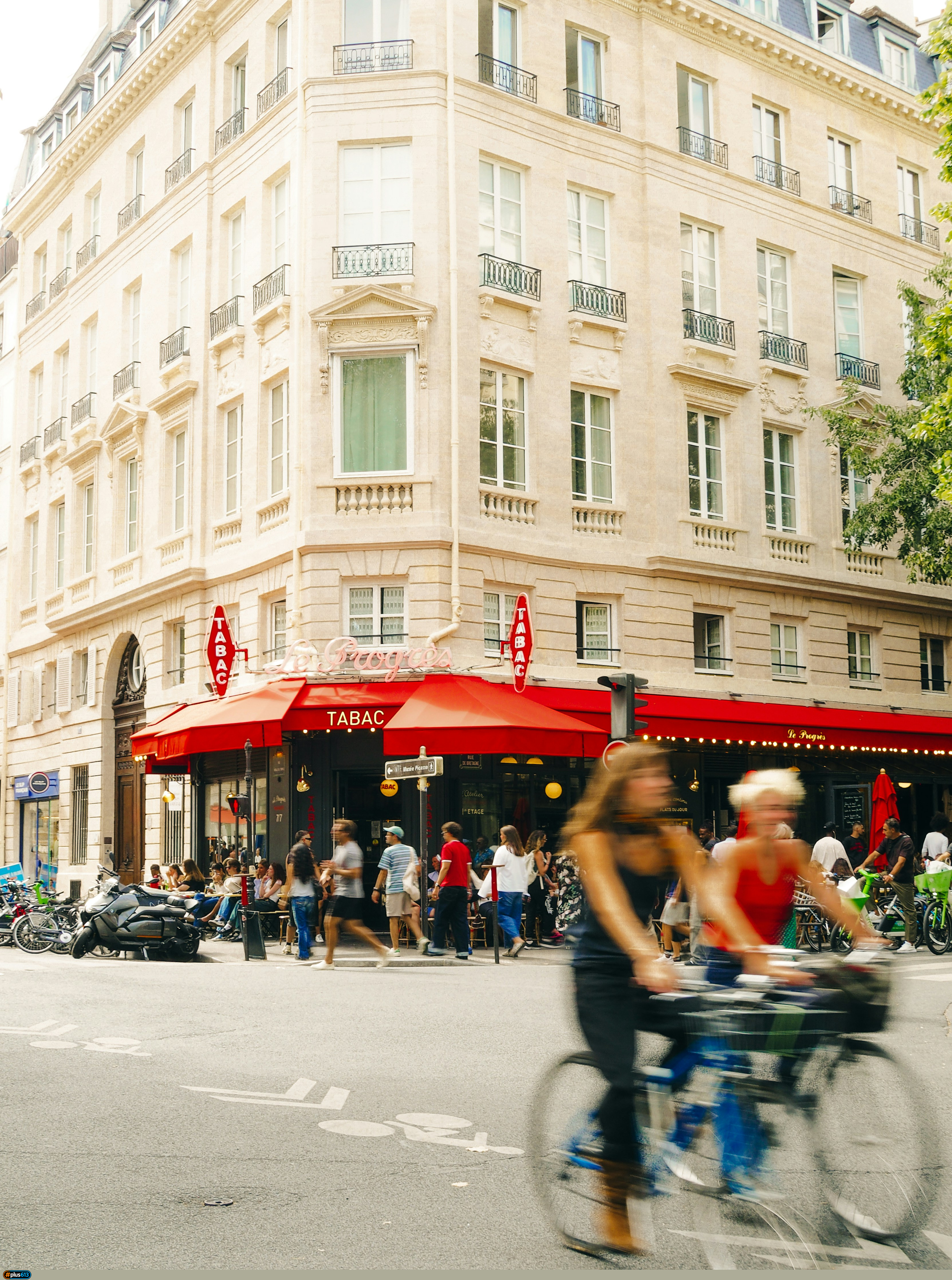 People cycling and walking past a corner cafe with red awnings in Paris by Lens by Benji
