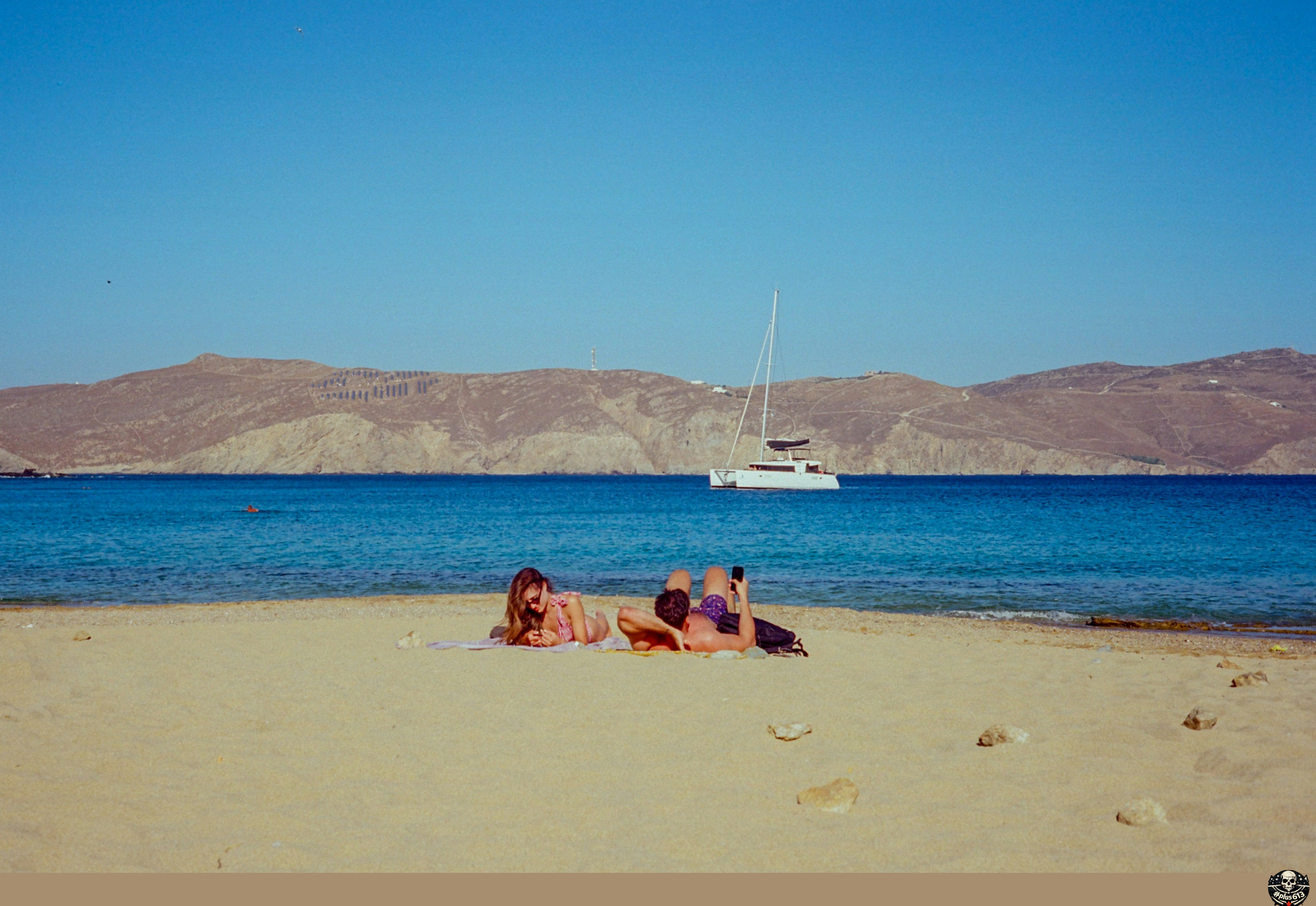 Two people sunbathing on a sandy beach with a sailboat by Jean Carlo Emer