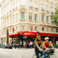 People cycling and walking past a corner cafe with red awnings in Paris by Lens by Benji